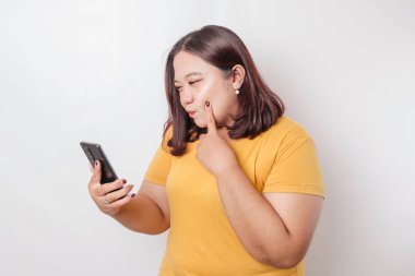 Telefonu izole edilmiş beyaz arka planda tutarken sarı gömlekli, iri yapılı Asyalı bir kadının kızgın ve kızgın yüzü..