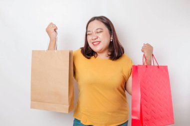 Mutlu, genç, Asyalı, iri yarı bir kadın sarı gömlek giyiyor ve elinde hediye kutusu ve alışveriş çantası tutuyor..