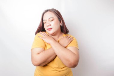 Genç, güzel, Asyalı, iri yarı, sarı tişörtlü, beyaz arka planda, mutlu, pozitif ve kendinden emin bir şekilde kendine sarılan bir kadın. Sevgi ve özgüven.