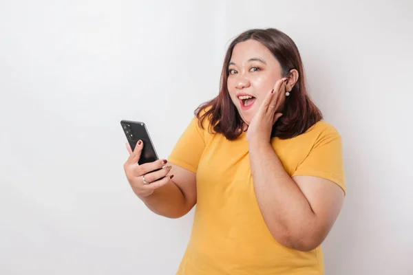 Zaskoczona Azjatycka Duża Kobieta Żółtej Koszulce Wskazującej Smartfona Odizolowana Białym — Zdjęcie stockowe