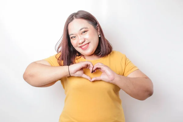 一位身穿黄色T恤的年轻貌美的亚洲大块头女子感到快乐 而一个浪漫的心形手势则表达了她的柔情 — 图库照片