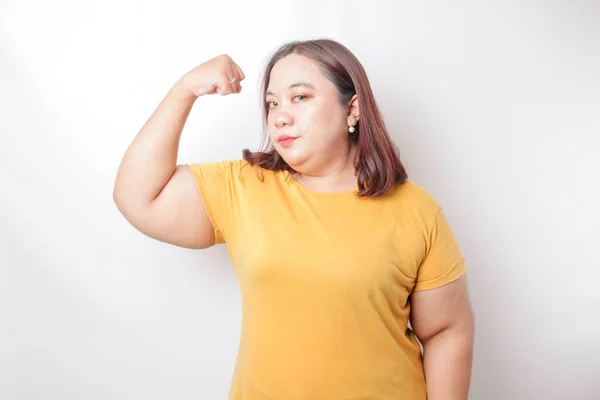 興奮アジアの大きなサイズの女性を身に着けています黄色のTシャツ彼女の腕や筋肉を持ち上げることによって強いジェスチャーを示す誇らしげに微笑む — ストック写真