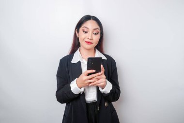 Mutlu bir Asyalı iş kadınının portresi gülümsüyor ve elinde akıllı telefonuyla beyaz bir arka planla izole edilmiş siyah bir takım elbise giyiyor.
