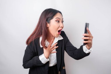 Siyah takım elbiseli Asyalı bir iş kadını elinde akıllı telefonuyla beyaz arka planda izole bir şekilde beklemekte.