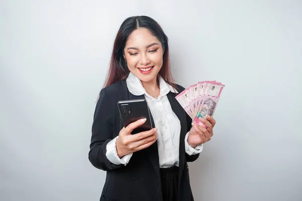一位快乐的年轻女商人身穿黑色西服 手里拿着手机和钱 用印尼盾和白色背景隔开 — 图库照片