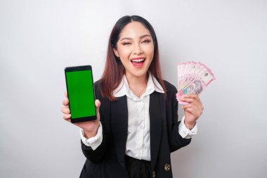 Genç ve mutlu bir iş kadını siyah takım elbise giyiyor. Telefonunu ve parasını Endonezya rupiasında gösteriyor. Beyaz arka planda izole edilmiş.