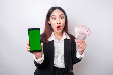 Siyah takım elbiseli Asyalı bir iş kadını Endonezya Rupia 'sında beyaz arka planla izole edilmiş akıllı telefonunu ve parasını gösteriyor.
