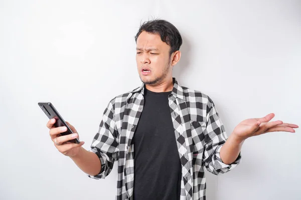 不満そうな若いアジアの男は 彼の携帯電話を保持するタータンシャツイライラ顔の表情を身に着けて不満そうに見えます — ストック写真