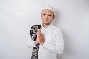 Omzunda dua halısıyla gülümseyen Asyalı genç Müslüman adam, beyaz arka planda izole edilmiş geleneksel selamlama hareketi yapıyor.