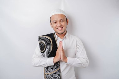 Omzunda dua halısıyla gülümseyen Asyalı genç Müslüman adam, beyaz arka planda izole edilmiş geleneksel selamlama hareketi yapıyor.