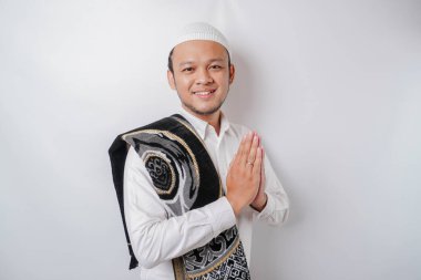 Omzunda dua halısıyla gülümseyen Asyalı genç Müslüman adam, beyaz arka planda izole edilmiş geleneksel selamlama hareketi yapıyor.