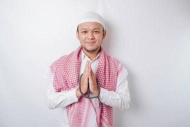 Gülümseyen Asyalı genç Müslüman adam, beyaz arka planda izole edilmiş geleneksel selamlaşmaları işaret ediyor.