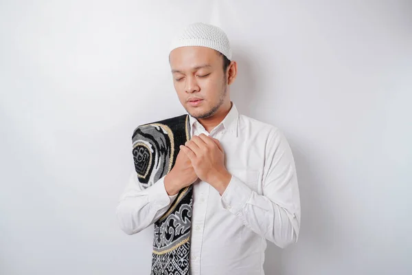 Happy handsome Asian Muslim man with prayer rug on his shoulder is praying to God.