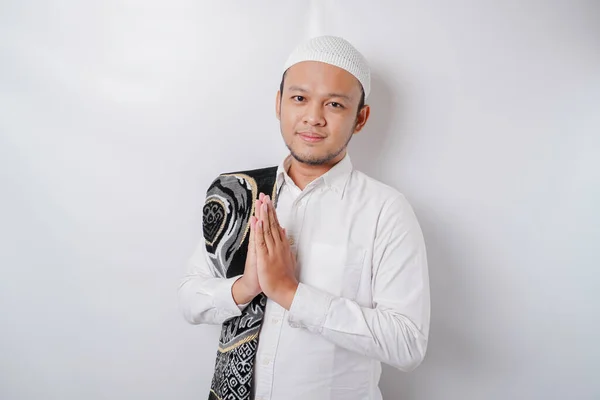 stock image Smiling young Asian Muslim man with prayer rug on his shoulder, gesturing traditional greeting isolated over white background