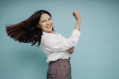 Kollarını ve kaslarını gururla kaldırarak güçlü bir hareket gösteren beyaz gömlek giyen heyecanlı Asyalı kadın.