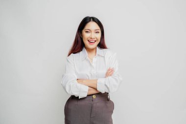 Beyaz tişörtlü, kollarını kavuşturup beyaz arka planda izole edilmiş kameraya bakan kendinden emin gülümseyen Asyalı bir kadının portresi.