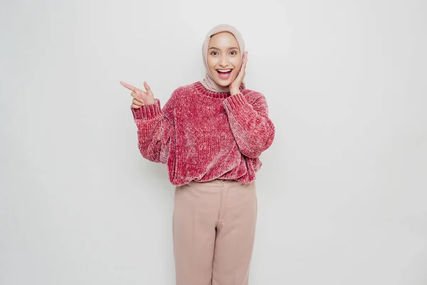 兴奋的亚洲穆斯林女人穿着粉色毛衣 头戴头巾 指着旁边被白色背景隔开的抄袭空间 — 图库照片