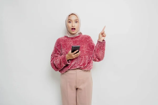 ピンクのセーターとヒジャーブを着た衝撃的なアジアのイスラム教徒の女性は 彼女の携帯電話を保持しながら 彼女の上にコピースペースを指して 白い背景によって隔離 — ストック写真