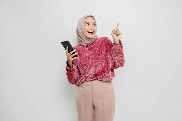 彼女の携帯電話を保持しながら 彼女の上にコピースペースを指してピンクのセーターとヒジャーブを身に着けている興奮したアジアの女性 白い背景によって隔離 — ストック写真