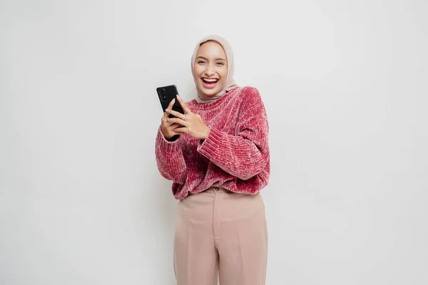 ピンクのセーターとヒジャーブを着た幸せなアジアの女性の肖像画 白い背景によって隔離された彼女の携帯電話を保持 — ストック写真