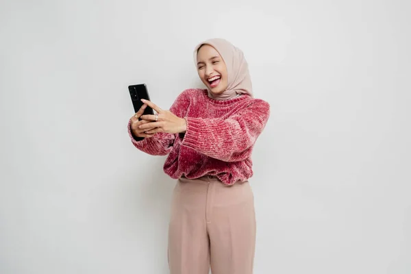 ピンクのセーターとヒジャーブを着た幸せなアジアの女性の肖像画 白い背景によって隔離された彼女の携帯電話を保持 — ストック写真