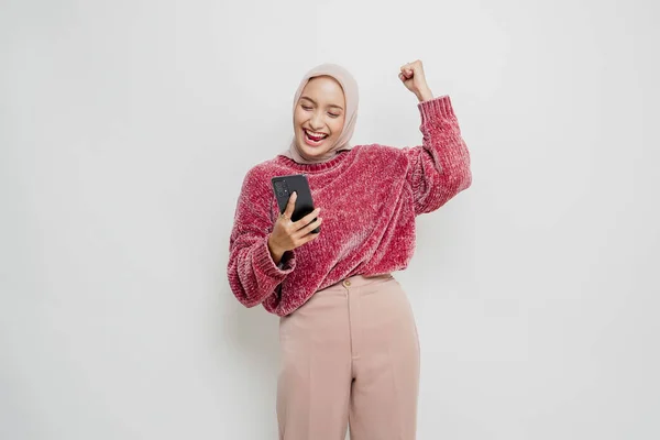 一位年轻的亚洲穆斯林妇女 穿着粉色毛衣 头戴头巾 手持被白色背景隔开的智能手机 表情快乐而成功 — 图库照片