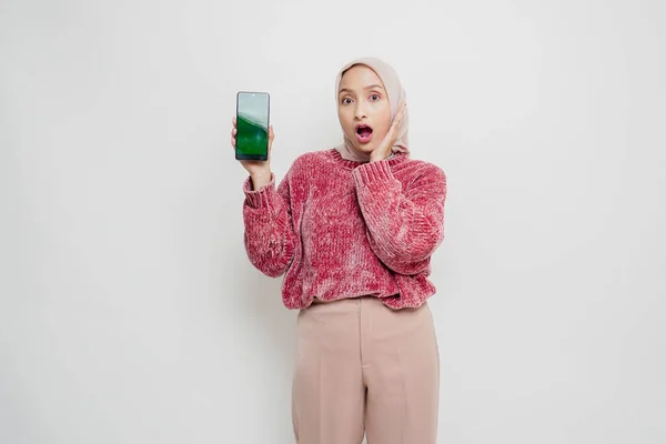 ピンクのセーターとヒジャーブを着たアジア系ムスリム女性が 白い背景によって隔離されたスマートフォンのコピースペースを見せている — ストック写真