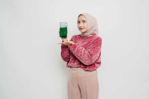 ピンクのセーターとヒジャーブを着た興奮したアジア系イスラム教徒の女性は 彼女の携帯電話を保持しながら 彼女の横にあるコピースペースを指して 白い背景によって隔離 — ストック写真