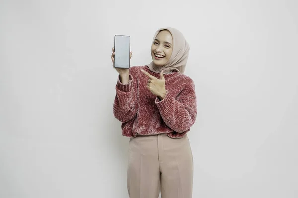 ピンクのセーターとヒジャーブを着た興奮したアジア系イスラム教徒の女性は 彼女の携帯電話を保持しながら 彼女の横にあるコピースペースを指して 白い背景によって隔離 — ストック写真