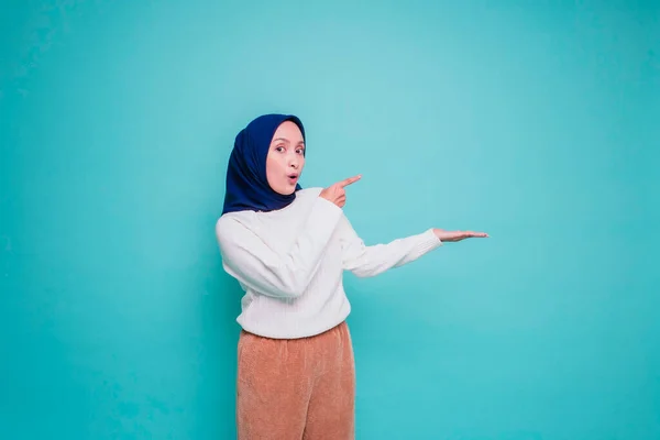 白いシャツとヒジャーブを着た衝撃的なアジア系イスラム教徒の女性は 青い背景によって隔離された 彼女の横のコピースペースを指して — ストック写真