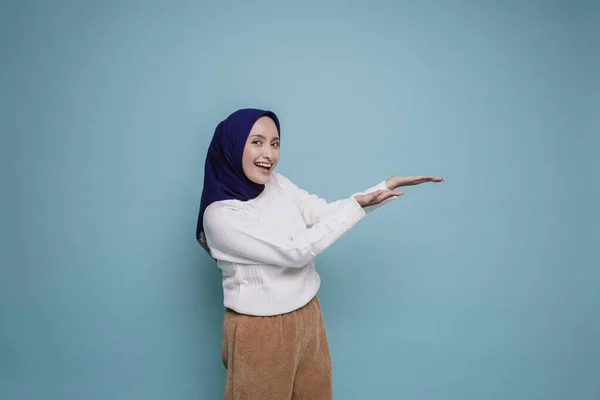 白いシャツとヒジャーブを着た興奮したアジア系イスラム教徒の女性が 青い背景によって隔離された彼女の横のコピースペースを指している — ストック写真