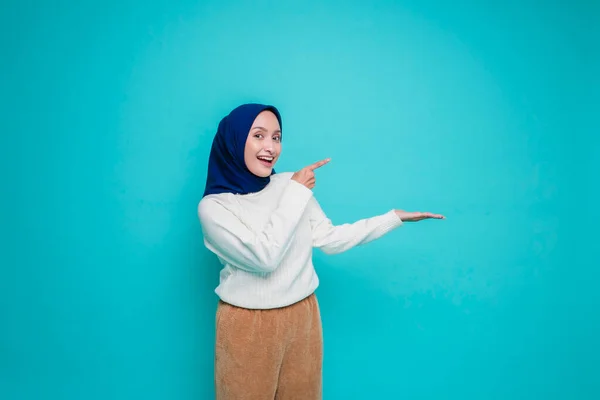 兴奋的亚洲穆斯林女人穿着白衬衫 头戴头巾 指着旁边被蓝色背景隔开的复制空间 — 图库照片