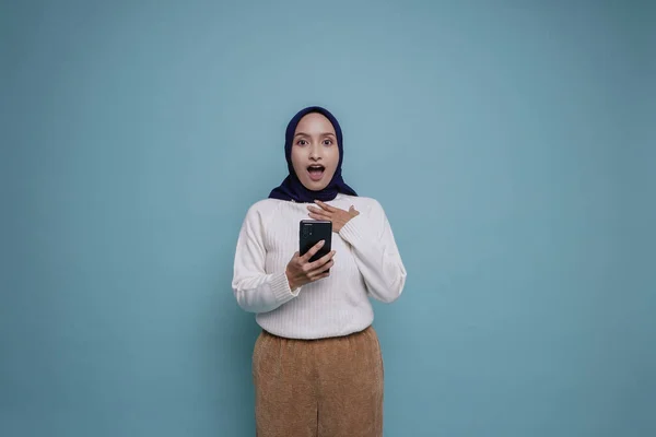 Zszokowana Azjatka Białej Koszuli Hidżabie Trzymająca Telefon Odizolowana Niebieskim Tłem — Zdjęcie stockowe