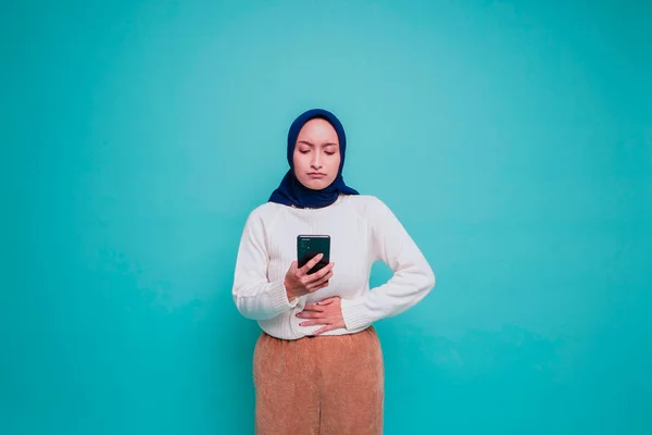 照片中 一个饥饿而兴奋的年轻穆斯林妇女拿着手机 想知道如何点蓝色背景的美味食品 — 图库照片