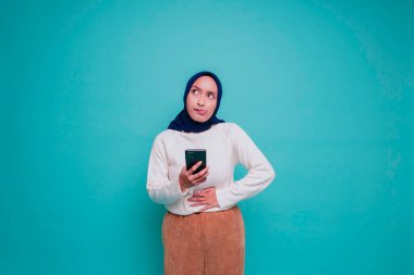 Aç, heyecanlı, genç bir müslüman kadının fotoğrafı. Elinde telefonu var ve mavi arka planda ne sipariş edeceğini merak ediyor.