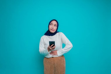 Aç, heyecanlı, genç bir müslüman kadının fotoğrafı. Elinde telefonu var ve mavi arka planda ne sipariş edeceğini merak ediyor.