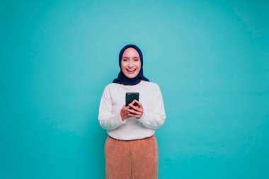 Beyaz gömlek ve tesettürlü mutlu bir Asyalı Müslüman kadının portresi, telefonunu tutuyor, mavi arka planla izole edilmiş.