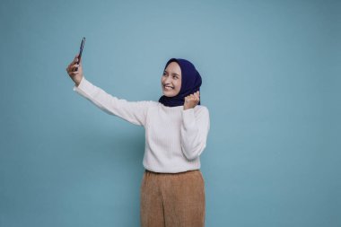 Mutlu yüzlü, beyaz gömlek ve tesettürlü ve elinde mavi arkaplan ile izole akıllı telefon tutan Asyalı genç bir Müslüman kadın.