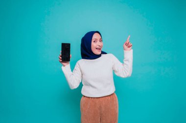 Telaşlı Asyalı Müslüman kadın beyaz gömlekli ve tesettürlü. Üstündeki fotokopi alanını işaret ediyor ve mavi arkaplan ile izole edilmiş telefon ekranını gösteriyor.