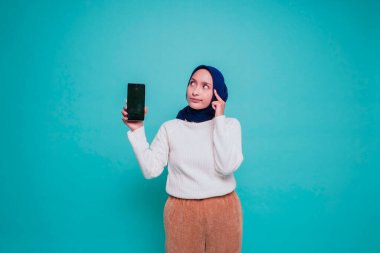 Düşünceli genç Müslüman bir kadın beyaz gömlek giyip çenesini tutarken telefon ekranında mavi arka planla izole edilmiş fotokopi odasını gösteriyor.