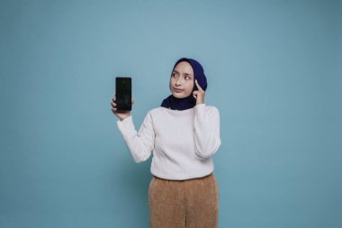 Düşünceli genç Müslüman bir kadın beyaz gömlek giyip çenesini tutarken telefon ekranında mavi arka planla izole edilmiş fotokopi odasını gösteriyor.