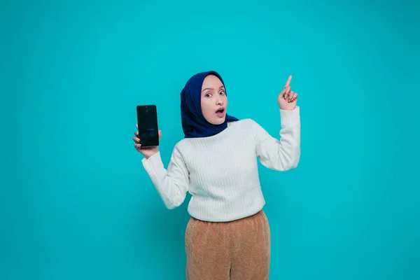 白いシャツとヒジャーブを着た興奮したアジア系イスラム教徒の女性が彼女の上のコピースペースを指して電話画面を見せ 青い背景によって隔離された — ストック写真