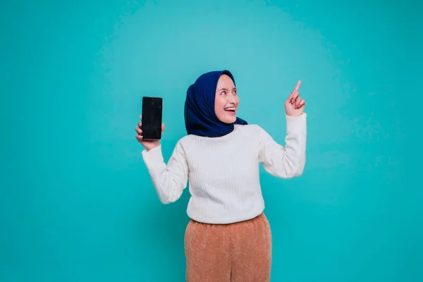 stock image Excited Asian Muslim woman wearing white shirt and hijab pointing at the copy space on top of her and showing phone screen, isolated by blue background