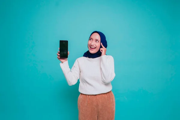 思慮深い若いイスラム教徒の女性は白いシャツを着ていると彼女の顎を保持しながら 彼女の携帯電話の画面にコピースペースを示す青の背景によって隔離 — ストック写真