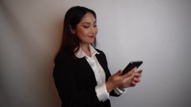 Asyalı genç bir iş kadını mutlu ve başarılı bir ifadeyle siyah takım elbise giyiyor ve elinde beyaz arka planla izole edilmiş akıllı bir telefon tutuyor.