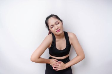 Spor kıyafetleri giyen hasta bir kadın elini karnına koyup beyaz arka planda acı çekiyor.