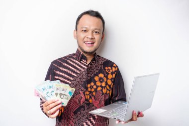 Mutlu bir Asyalı genç adam batik gömlek giyiyor, elinde Endonezya rupiasında beyaz arka planla izole edilmiş bir laptop ve para var.