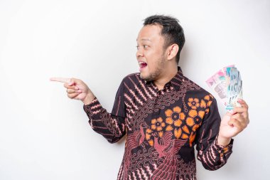 Mutlu bir Asyalı genç adam batik gömlek giyiyor, yanındaki fotokopi alanını işaret ediyor, Endonezya rupiasında beyaz arka planla izole edilmiş para tutuyor.