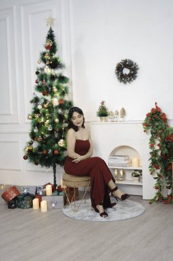 Portrait of pretty young girl cozy sit down, smiling wear red gown in decorated Christmas living room indoors