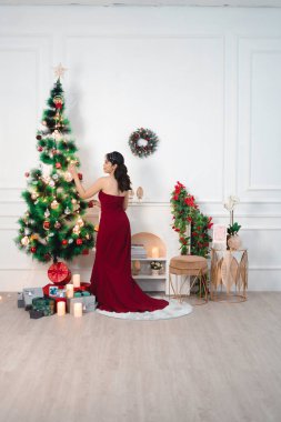 Portrait of pretty young girl decorating Christmas tree, smiling wearing red gown in decorated Christmas living room indoors
